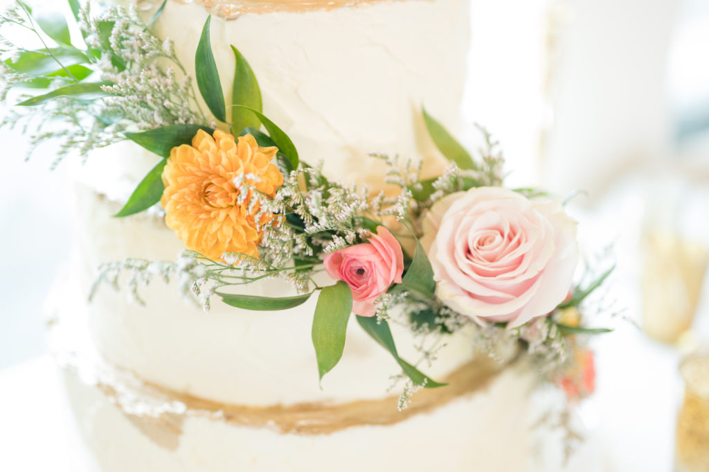 White wedding cake with gold accents is surrounded by pink and orange flowers, as well as greenery. 
