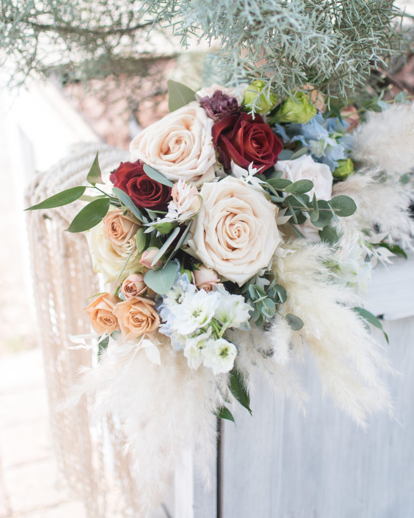 A Wedding bouquet wrapped in macramé features light pink, red and orange roses with eucalyptus and an assortment of pastel flowers for a fall wedding color palette