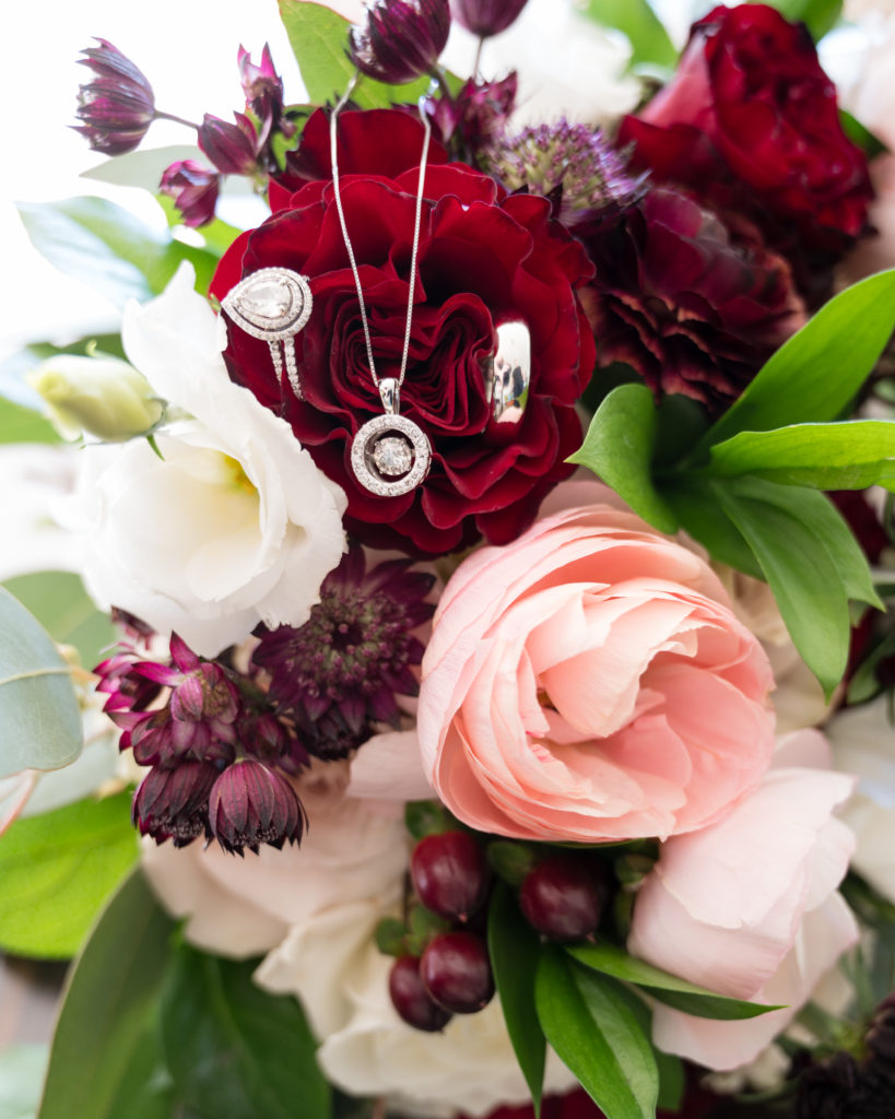 A wedding bouquet displays the couples wedding rings and brides necklace with a fall wedding color palette of pink and burgundy