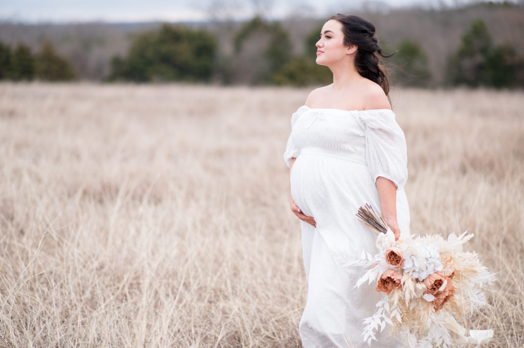 Maternity session with a sweet mama holding her belly in a white dress as she looks into the wind and holds a bouquet of dried flowers.