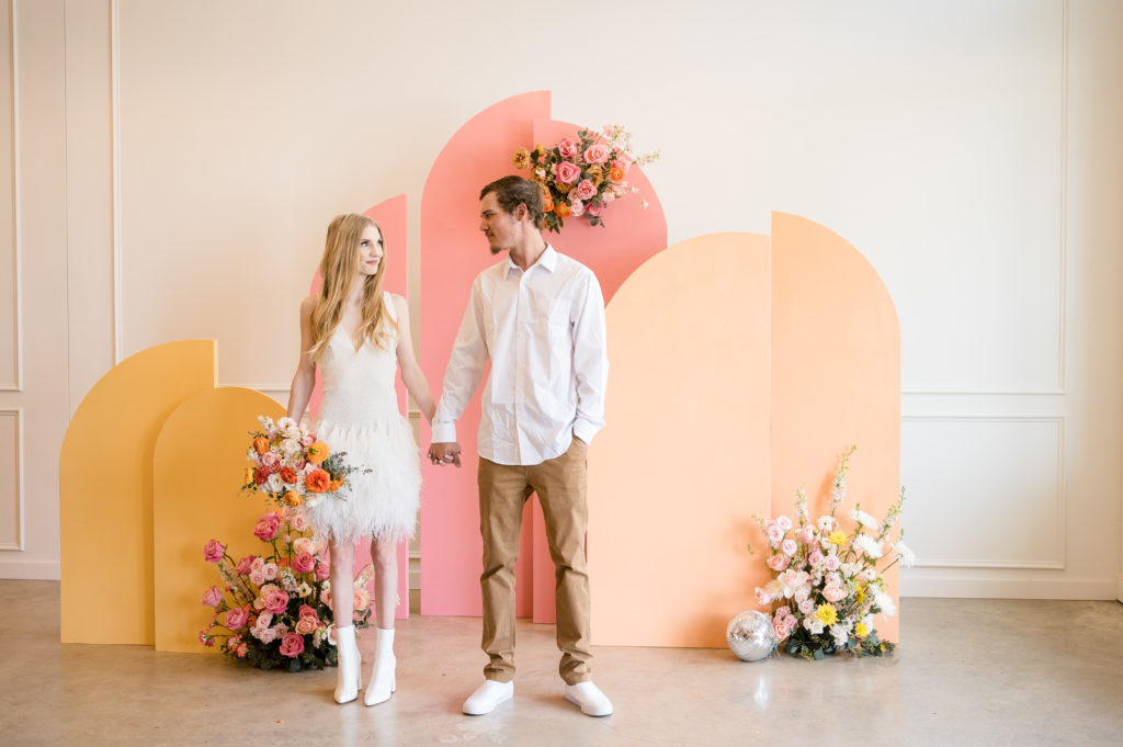 Couple in a retro wedding flapper dress stand in front of a colorful backdrop full of flowers
