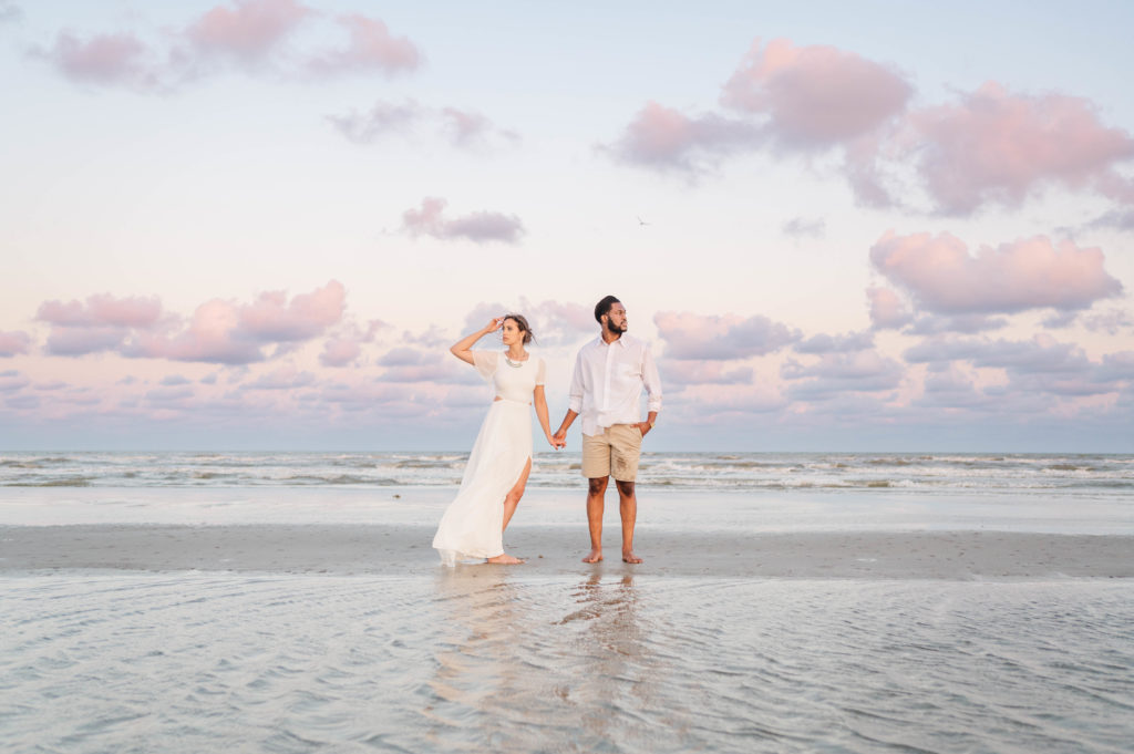Bride and groom stand on a sand bank in the ocean with the background of a pink sunset in Galveston Texas.