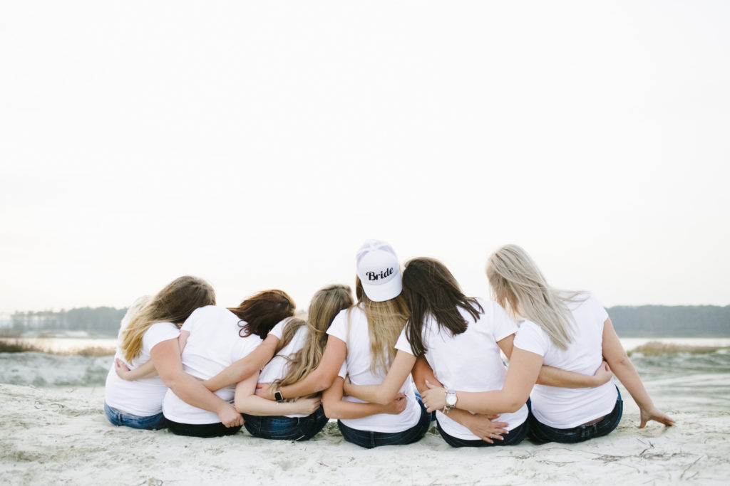 Group of girls sit arms around each other on the beach, the center wears a hat that says "Bride" 
